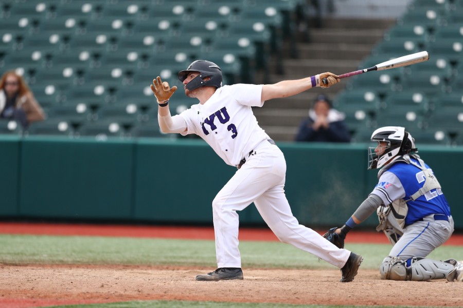 Captured in motion on the baseball field, this picture shows a baseball player dressed in a white baseball jersey that has dark purple “NYU 3” printed on the front, a pair of white pants, a black helmet, black sneakers and a pair of light brown gloves. His left hand is holding a steel baseball bat. Kneeling on the right side of the image next to him is another player dressed in a blue and gray uniform.