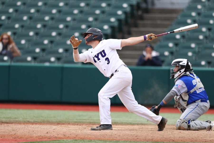 Shot+in+motion+on+baseball+field%2C+on+picture+shows+a+baseball+player+dressed+in+white+baseball+shirt+in+dark+purple+%E2% 80%9CNYU+3 %E2%80%9D+print+on+front%2C+a+pair+white+pants%2C+a+black+helmet%2C+black+sneakers+and+a+pair+light+brown+gloves.+His +left+hand+holds+steel+baseball+bat+bat.+Stands+on+knees+on+right+side+images+near+to+he+it+is+another+player+dressed+in+blue+and+ gray + shape.