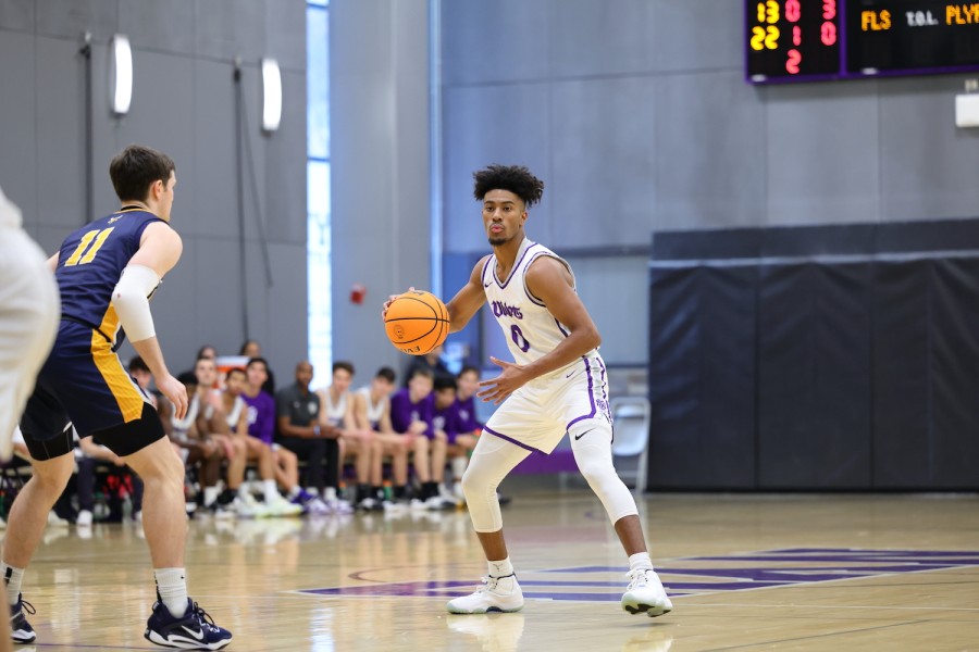 An N.Y.U. basketball player stands in the middle of a basketball court. He is dribbling a basketball. A member of the opposing team guards in front. In the background, a group of people watch the game from the stands.