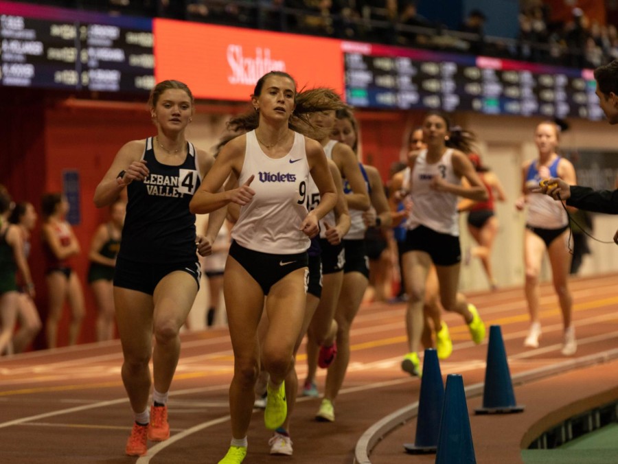 A group of around eight runners are pictured running on an indoor track, wearing athletic tank tops and shorts.