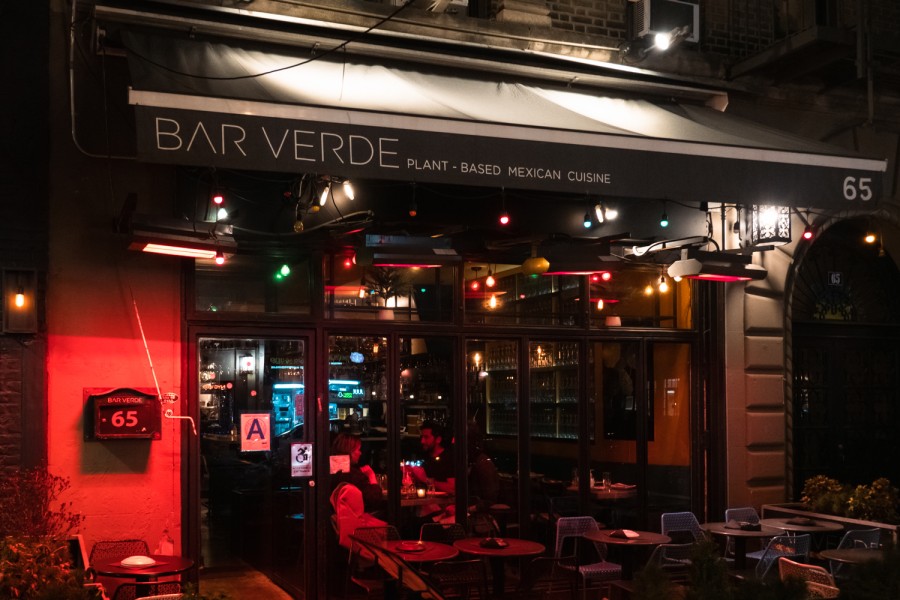 The storefront of a vegan restaurant named “Bar Verde.” The windows and entrance are lit up with red lights and there are two people dining inside.