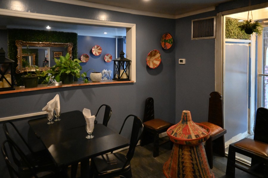 Photo of a dimly lit restaurant with a dark blue wall.  There is a black table with black chairs, a window-shaped hole in the wall with plants and candles inside, a mirror in the background, and a few more chairs against the walls in the foreground.