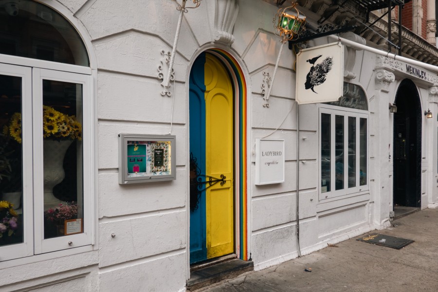 A bright, colorful, arched door is at the center of the restaurant Ladybird’s white exterior. A vase of sunflowers can be seen behind the window on the left side of the wall.