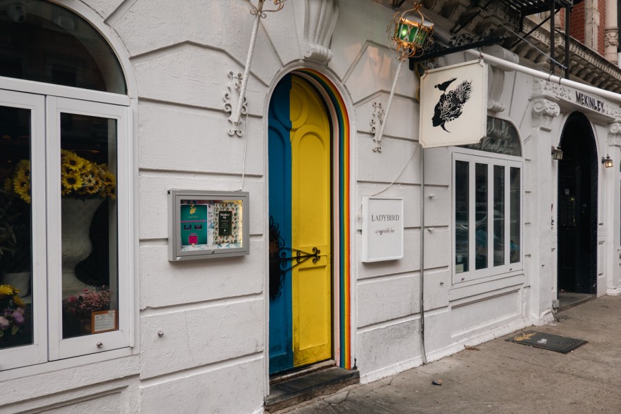 A bright colorful arched door is at the center of the white exterior of the Ladybird restaurant.  A vase of sunflowers can be seen outside the window on the left side of the wall.