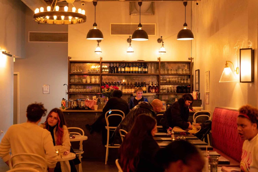 In the warmly lit vegan restaurant Modern Love, a woman is behind a bar and in front of a wooden wine cabinet. A room of customers occupies the foreground of the image.