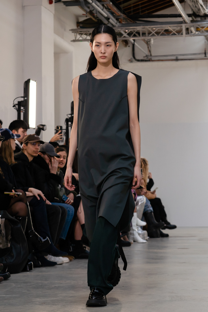 A model wearing a dark blue-green sleeveless dress with loose-fitted black pants walking down a runway.