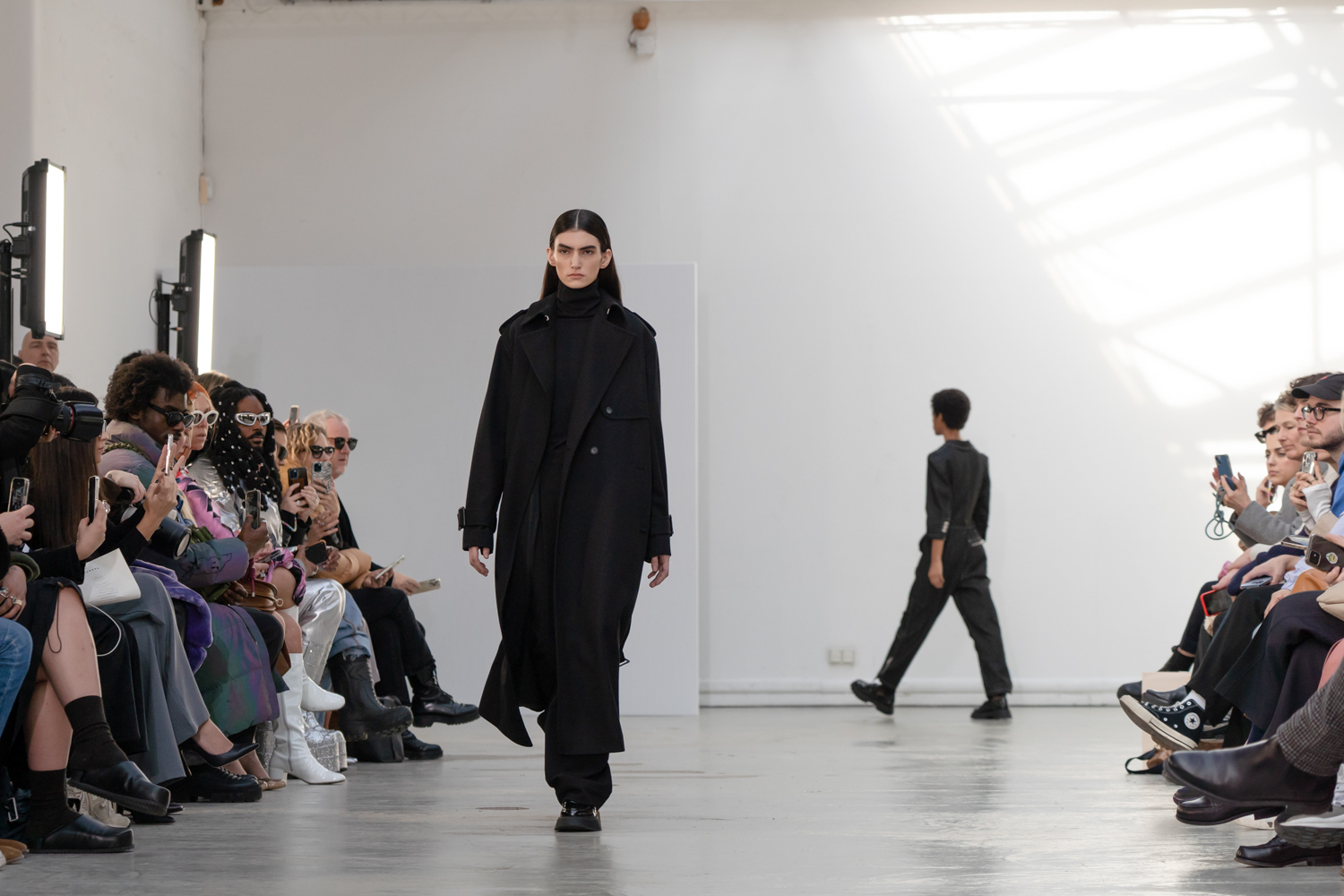 A model wearing a black turtleneck, trench coat, and trousers walking down a runway.
