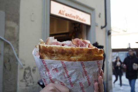 A hand holding a prosciutto sandwich, wrapped in paper.