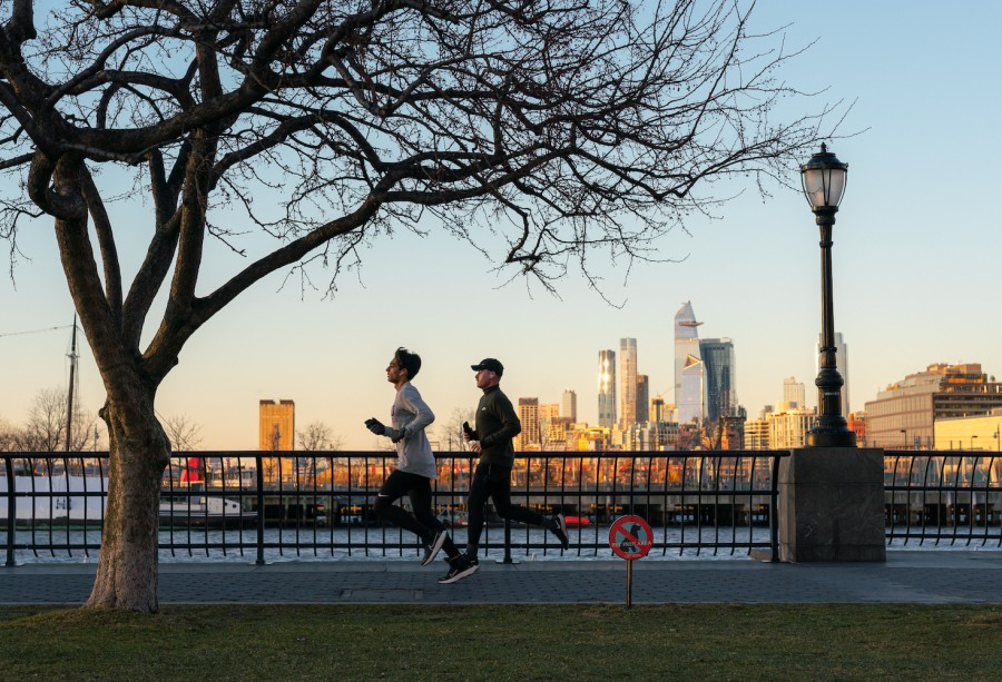 Two runners run across a pier on Hudson River. The skyscrapers at the Hudson Yards are further away in the background with the sun shining on them.