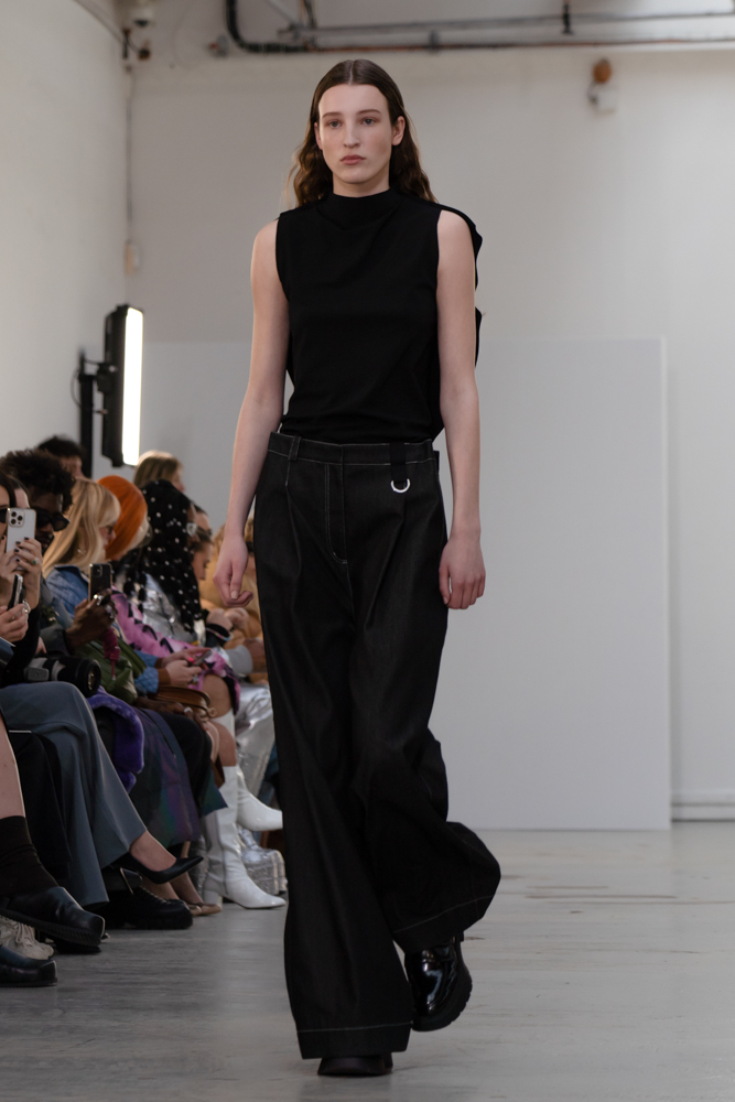 A model walking down a runway wearing a black mock-neck sleeveless top and baggy pants.