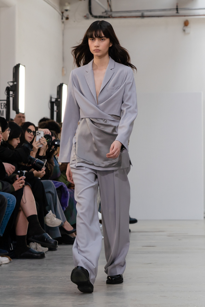 A model in a light gray wrap shirt and loose-fitted pants walking down a runway.