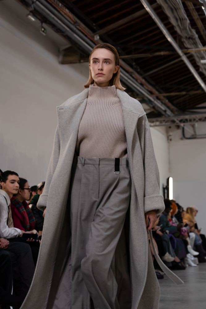 A model wearing a beige sweater, long gray coat and pleated trousers walking down a runway.