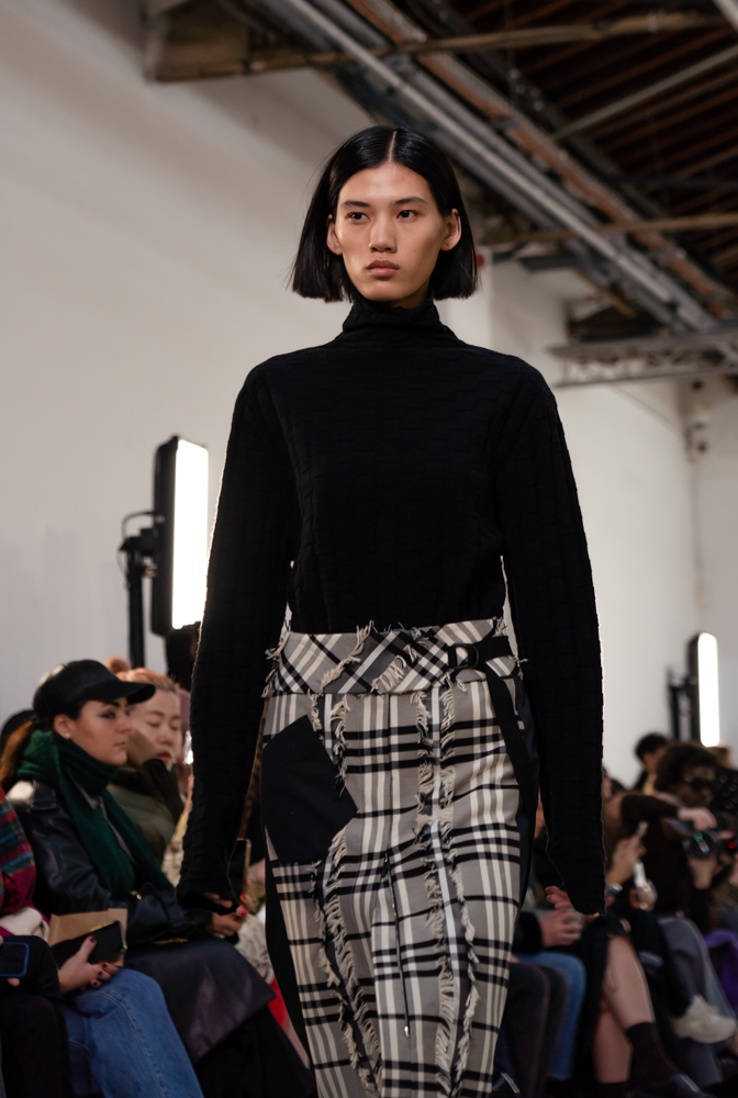 Close-up of a model walking down the runway in a black turtleneck and patched plaid skirt.