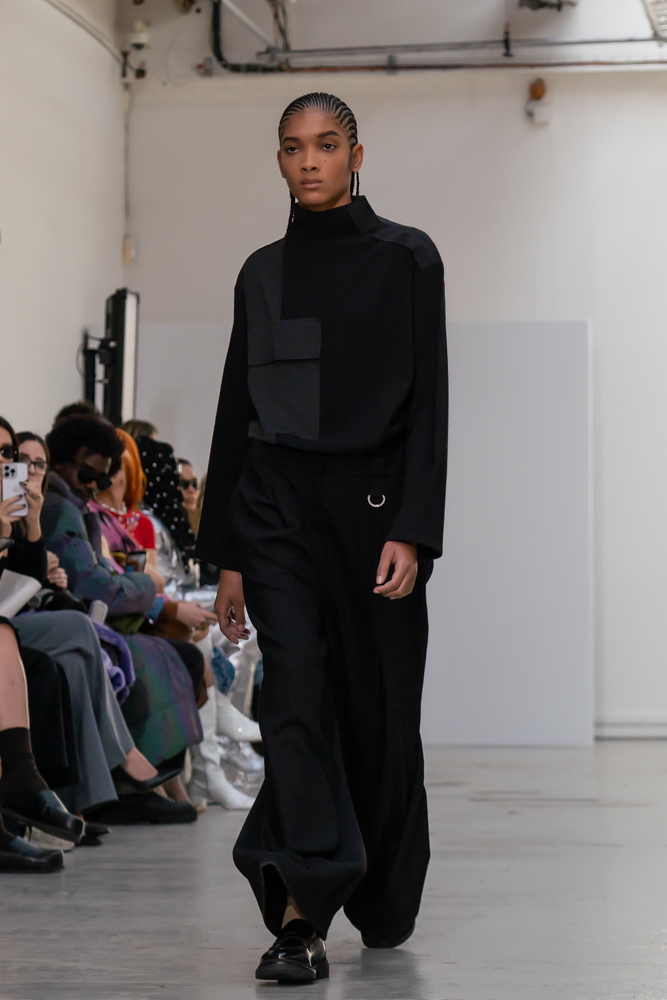 A model wearing a patched black and gray shirt with loose-fitting black trousers walking down the runway.
