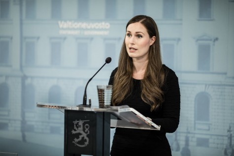 Sanna Marin, wearing a black dress, stands behind a glass podium which has a microphone attached to it. The podium has the emblem of the Finnish government on it. There is a blue wall behind her, with the logo of the Fnnish government printed on it.