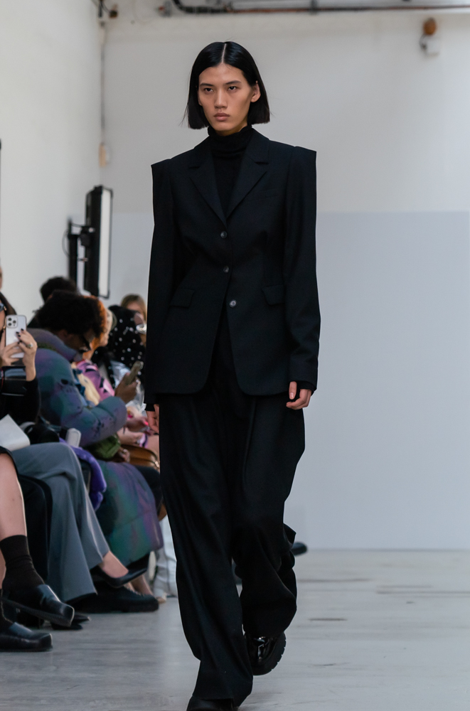 A model in a black turtleneck, shoulder-padded blazer, and trousers walking down the runway.