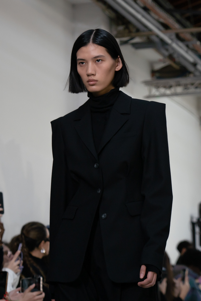 Close-up of a model walking in a black turtleneck, shoulder-padded blazer, and trousers.