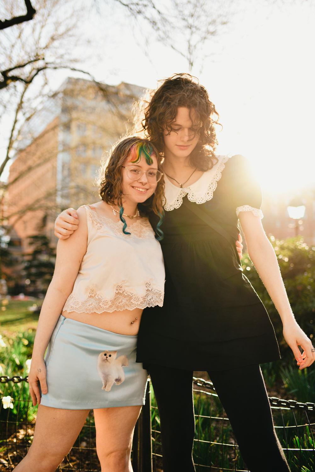 Ariel Rond and Saoirse Sowell stand together in a park with the sun shining on them. They have their arms around one another and smiling at the camera.