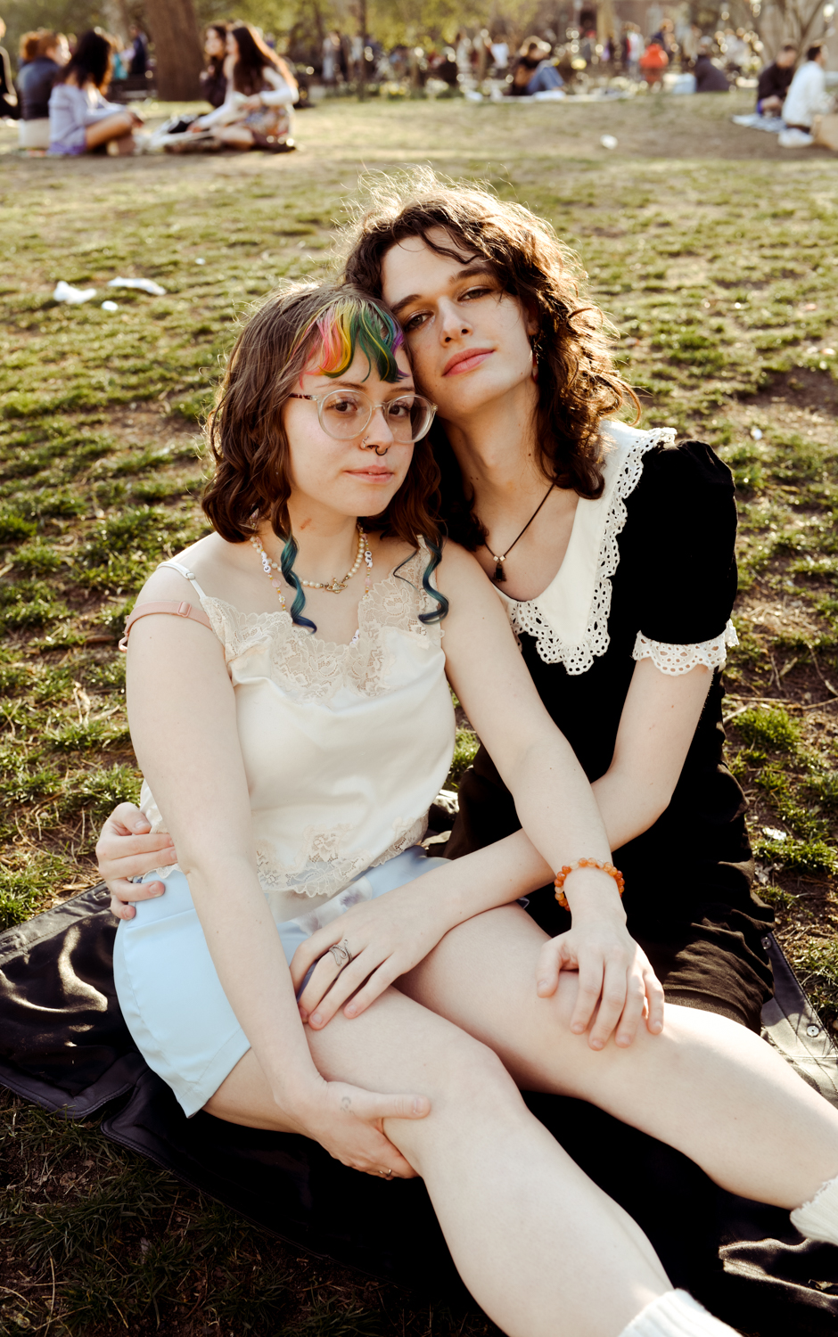 Saoirse Sowell and Ariel Rond sit together on the grass in a park with the sun shining on them.