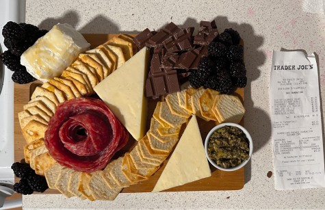 A charcuterie board consisting of cheese, croutons, salami, blackberries, chocolate and a bowl of pesto is assembled on a wooden cutting board.  Next to the board is a receipt from Trader Joe's with the prices of every item on the board.