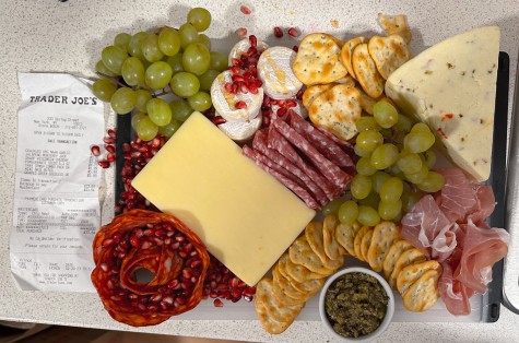 A charcuterie board is assembled, consisting of crackers, cheese, grapes, pomegranate seeds, various cured meats and a bowl of pesto.