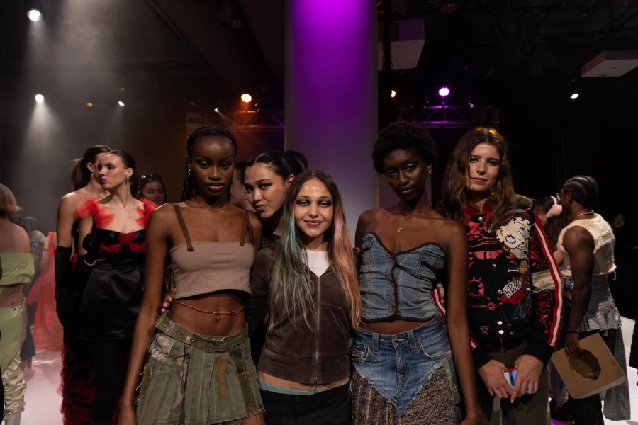 A group of models stand together for a photo on the runway. Behind them is a stream of purple light.