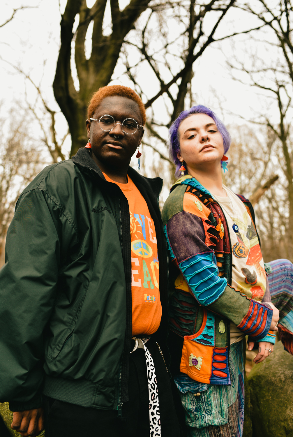 Dee Oglesby and Jinx Sage stand together in Prospect Park. They are looking down into the camera. Oglesby, who has orange hair, is wearing black pants, an orange t-shirt, and a green jacket, and has a nose ring. Sage, who has purple hair, is wearing a bright-colored patchwork jacket and patchwork pants.