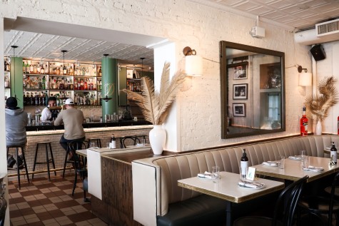 The interior of a well-lit restaurant with white interior walls. There are tables and seats in the front and a bar at the back.