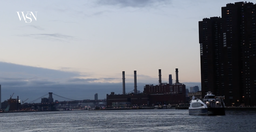 A ferry runs on the East River with the Williamsburg Bridge and many buildings in the background.