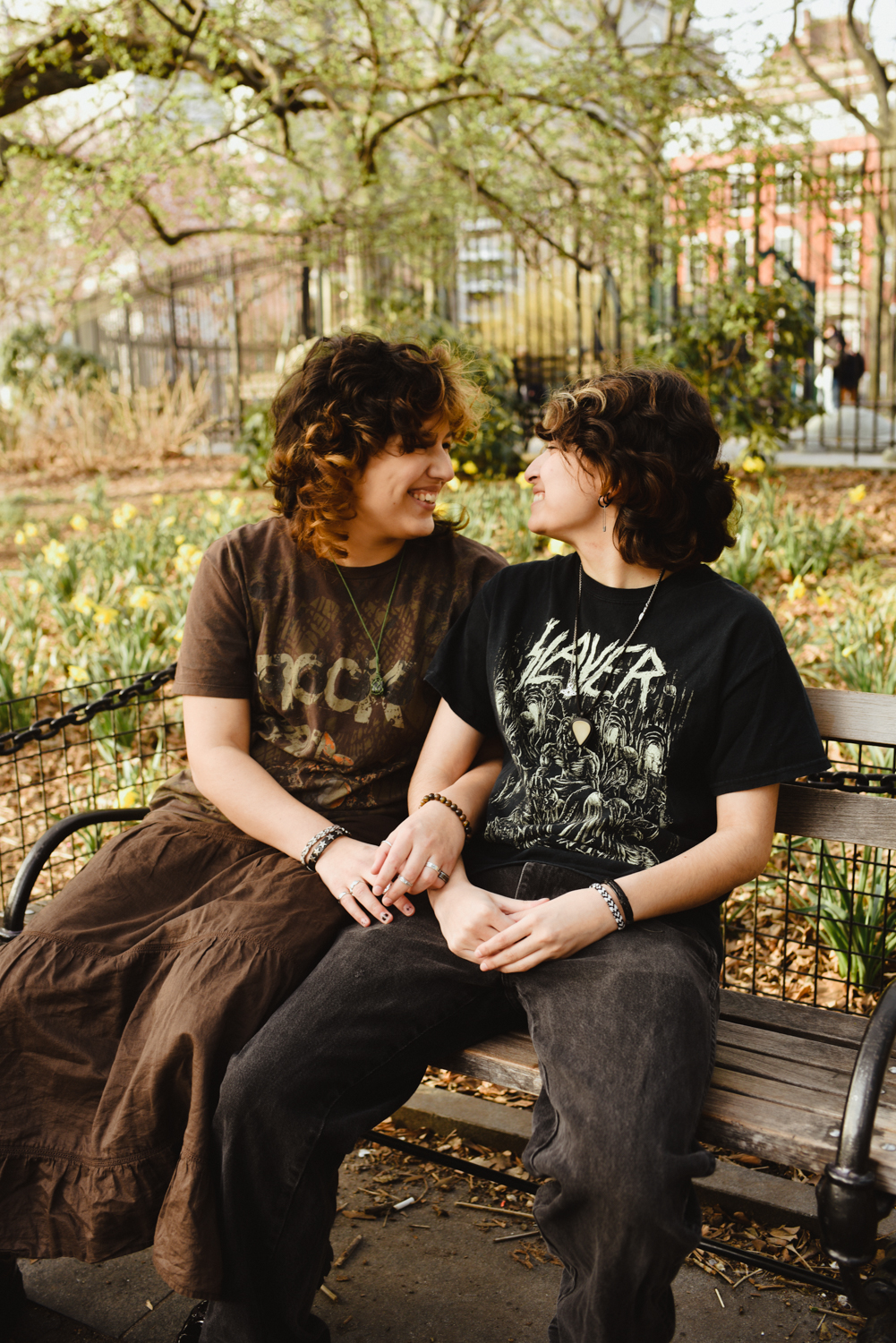 Gigi Portilla and Miche Fernandez sit together on a bench under a tree in Washington Square Park. They are looking at each other and laughing.