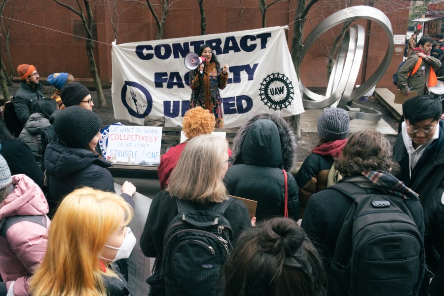 A speaker holding a white megaphone delivers a speech in front of rally attendees. Behind the speaker is a white banner with blue text that reads “Contract Faculty United.”