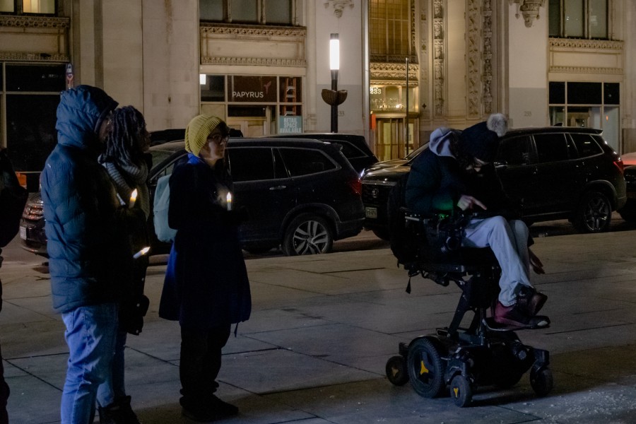 Four vigil attendees appear in the photo. The three on the left stand, each holding a candle. The one on the right sits in a powered wheelchair.