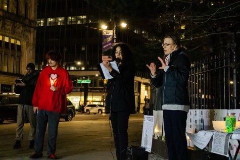 Four vigil attendees appear in the photo. The two on the right recite to the crowd from a script. An attendee on the left holds a candle in their hand, next to someone reading a prepared statement.