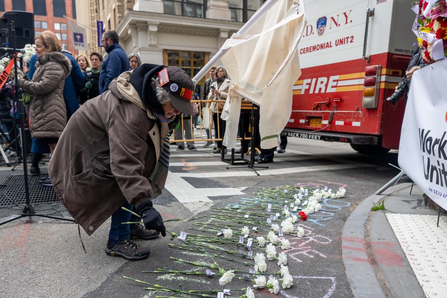 A+woman+in+a+brown+jacket+bends+down+and+lays+a+white+flower+on+the+pavement+among+other+flowers.+In+front+of+her+is+a+banner+and+a+wreath%2C+behind+it+is+a+fire+truck.