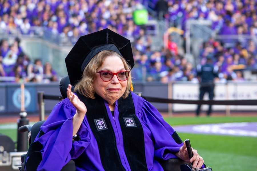 Judith Heumann wearing N.Y.U’s commencement robe and red-framed glasses waves to the crowd.