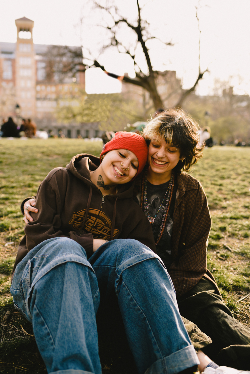 Koda Fraga and Leo Koulish sit together on the grass in Washington Square Park with the sun shining on them. They are holding each other and smiling at the camera.
