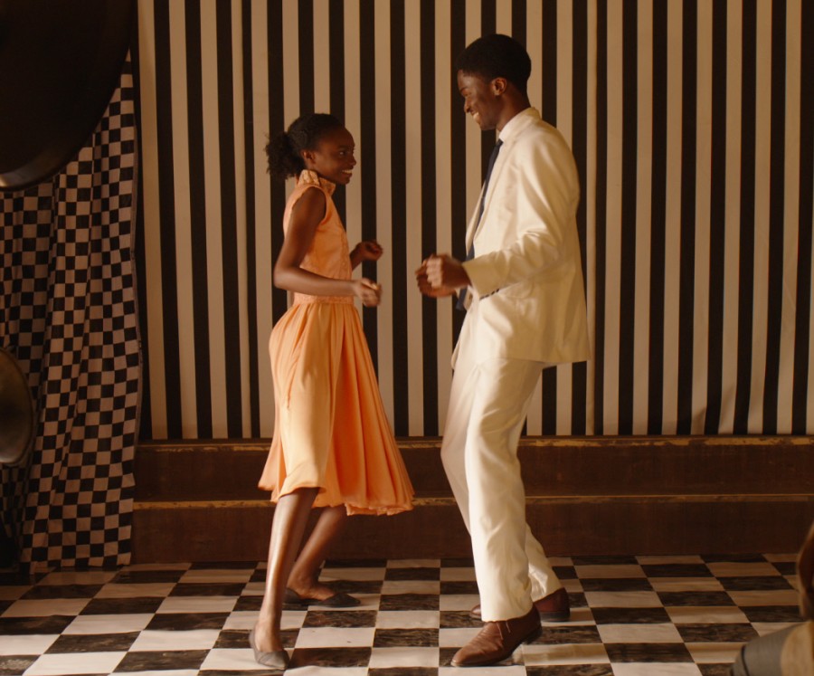 A couple, one person wearing an orange dress and the other wearing a light tan suit, dance on a black-and-white tile floor.