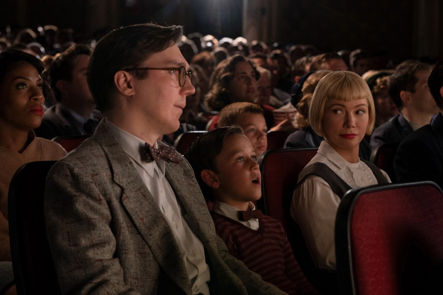 A family — including a mother with a blonde bob haircut who is wearing a white shirt and a black dress, a son, and a father wearing a gray, plaid three-piece suit — watches a movie in the theater.