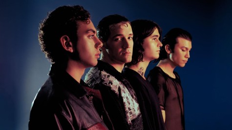 A photo of the four members of the band Quarters of Change standing in a line showing the sides of their bodies. They are wearing black clothes and are standing against a blurred blue background.