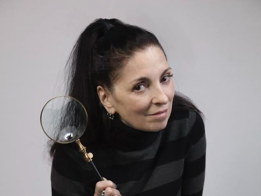 Musician Palmyra Delran wearing a black jacket while holding a magnifying glass in her hand. She sits in front of a white backdrop.