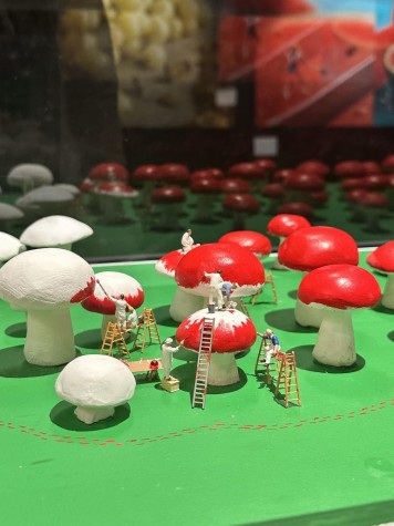 An exhibit with several miniature mushrooms with ladders carrying miniature human beings. This is in a display case with a green base.