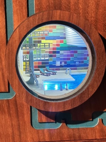 An exhibit with a circle on a wooden panel. Inside the circle is a miniaturized scene of a swimming pool with two lawn chairs in front of a rainbow tile background.