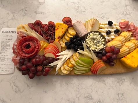 A charcuterie board consisting of crackers, cheese, blackberries, grapes, apple slices, various cured meats and madeleines is assembled on a wooden cutting board.  Nearby is a receipt from Trader Joe's with the prices for each ingredient.