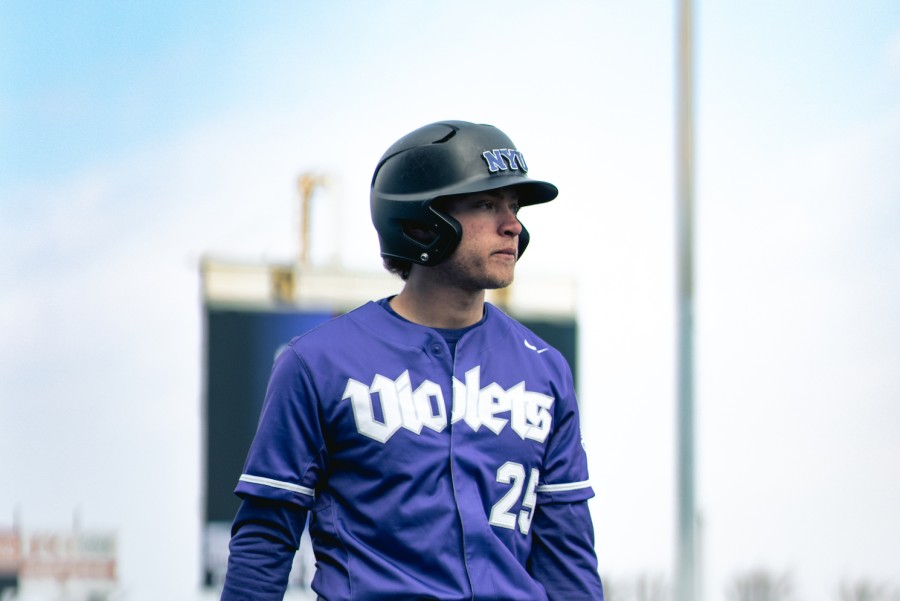A man in a purple uniform with the word “Violets” and the number 25 on it stands in front of a large display screen under a blue sky. He is wearing a black batting helmet which reads “N.Y.U.” in purple.