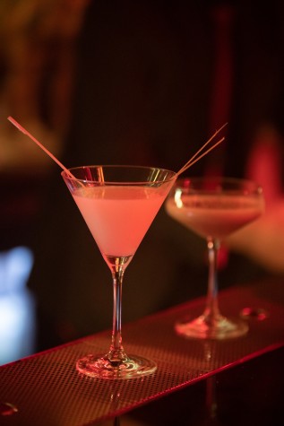 A white, opaque drink with strings of ginger on the side in a conical wine glass on a bartop, lit in red, atmospheric light.