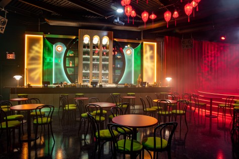 A wide interior shot of a dining area in the foreground and a bar lit in green and yellow light in the background. Red lanterns hang from the ceiling in the top right of the image. Bottles of liquor sit on a shelf above the bar.