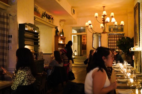 The interior of a restaurant at night with warm lighting and several customers inside. A chandelier is hanging from the ceiling and there are small candles placed on each table.