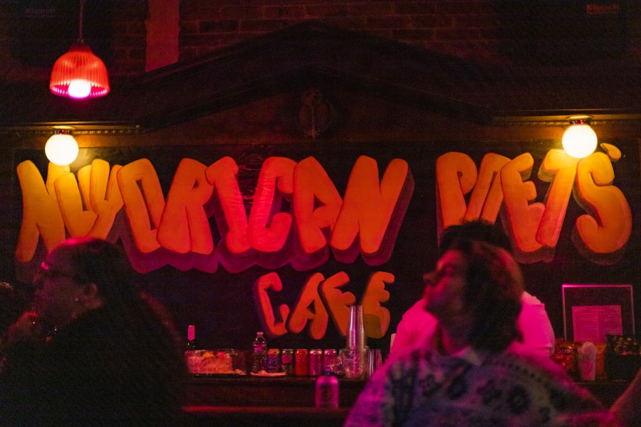 A bar counter with several people sitting next to it. There is painted text reading “Nuyorican Poets’ Cafe” in all capital letters on the wall behind the counter.