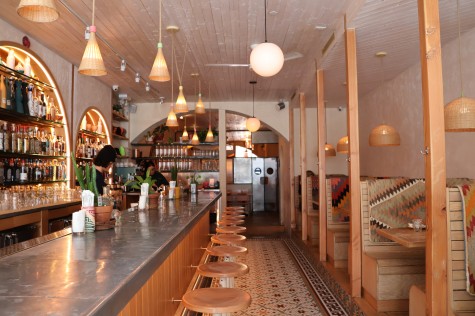 The wooden interior of a restaurant. Hanging from the ceiling are pendant lights. To the right is a seating area. To the left is a bar.