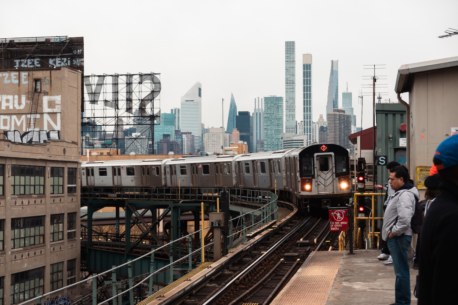 The 7 train arrives at the outdoor platform with the midtown Manhattan skyline in the background. There are people waiting on the platform.